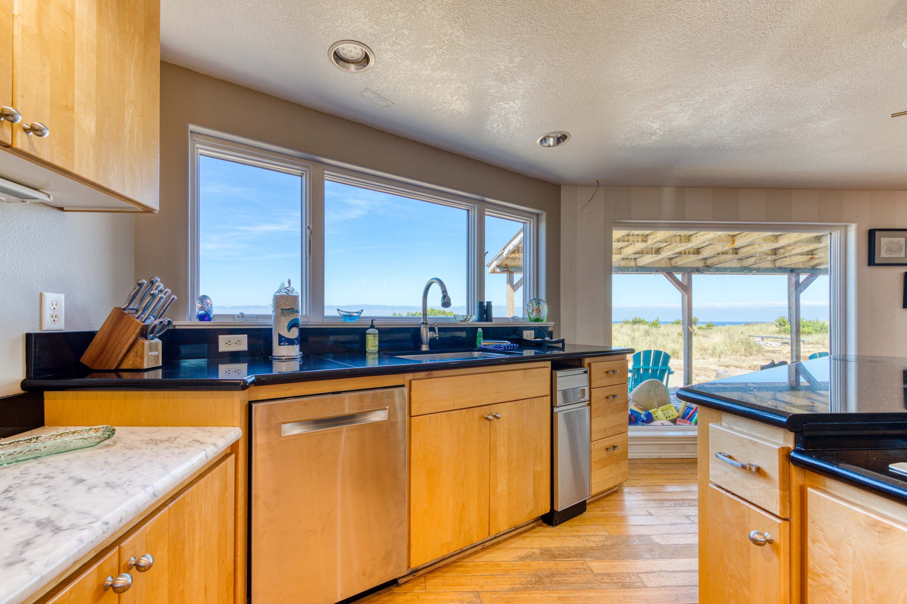 A modern kitchen with wooden cabinets, a black countertop, and a view of the beach through large windows.