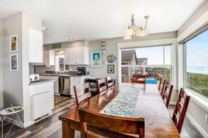 Cozinha moderna com armários brancos, área de jantar com uma mesa de madeira comprida e vista para a praia através de grandes janelas.