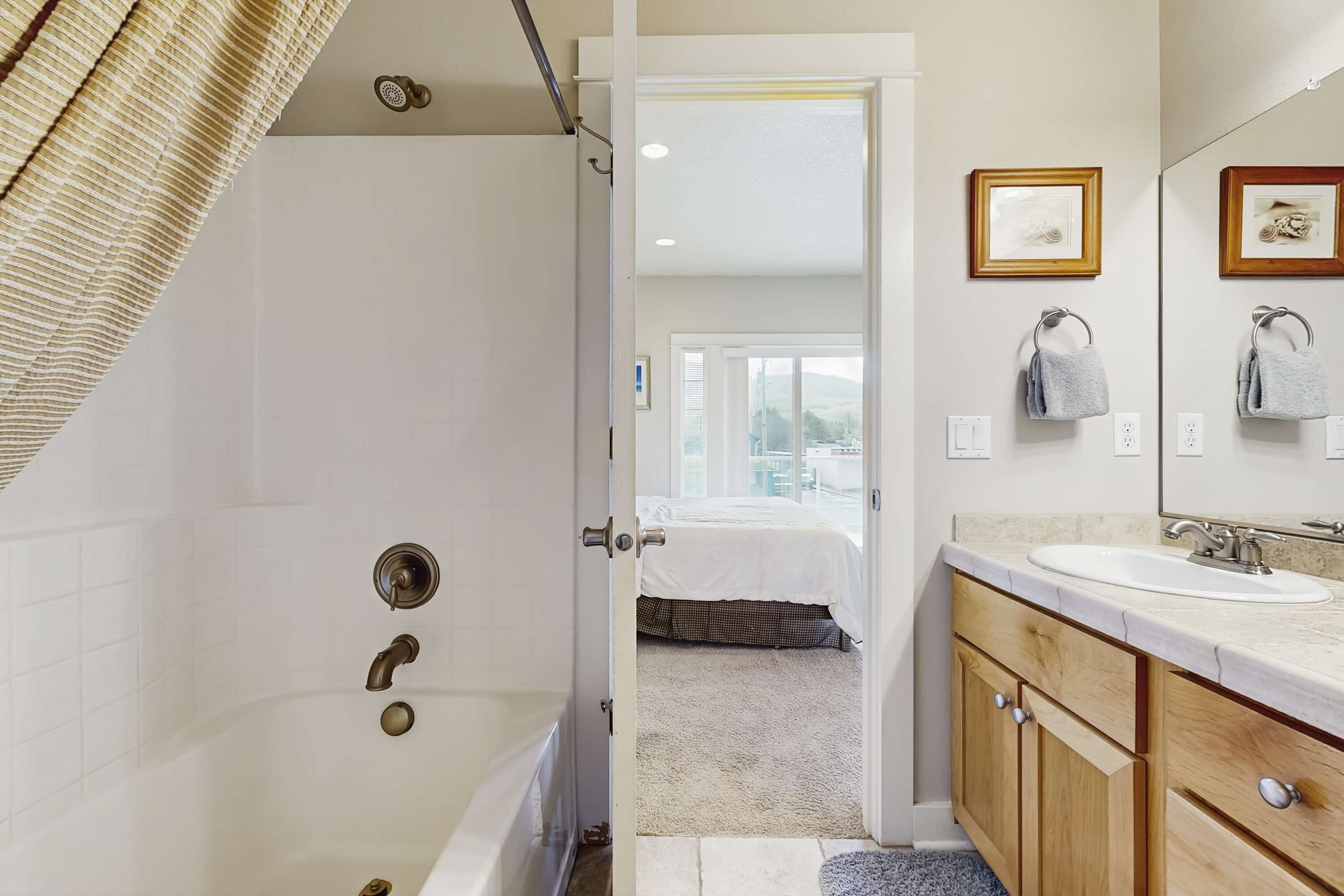 A modern bathroom with a bathtub, a sink, and a mirror. There is a door leading to a bedroom with a bed and a window.