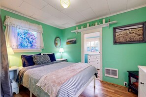 A bedroom with a bed, bedside tables, a lamp, a window with a floral curtain, a mirror, a shelf with decorative items, and a wall-mounted picture.