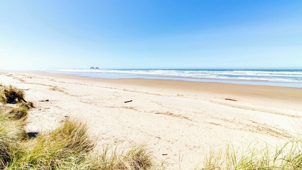A beach with sand and grass in the foreground, a clear sky, and a calm sea. - New House in Quiet Location with Modern Amenities near Beach Lake and Town (Rockaway Beach)