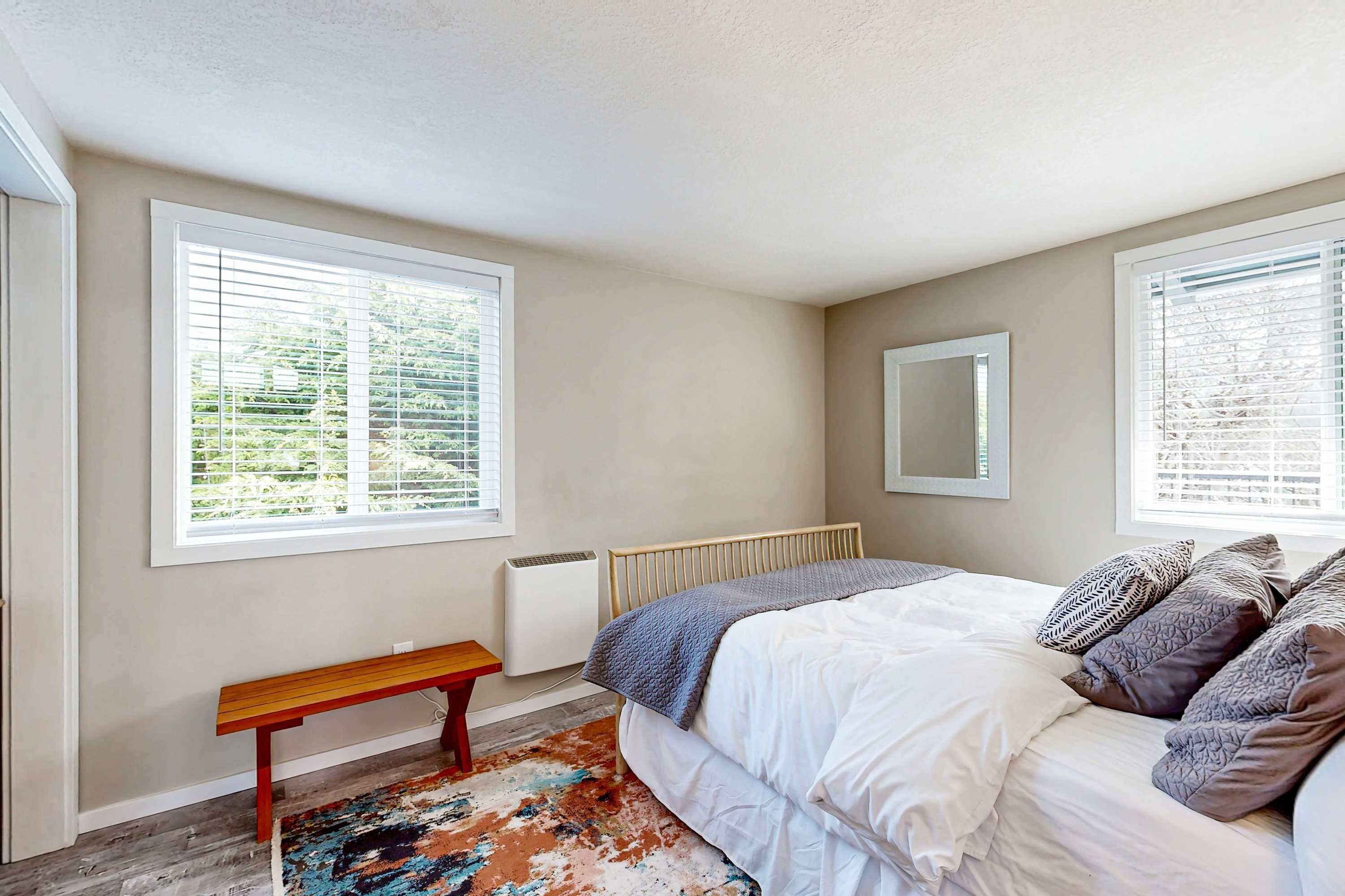 Quarto com cama, um banco, radiador e janelas amplas.