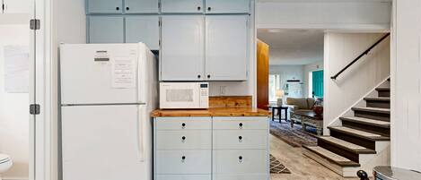 Une cuisine compacte équipée d’un réfrigérateur, d’un four à micro-ondes et de placards bleu clair. Un plan de travail en bois surmonte les placards. Un escalier avec une rampe métallique est visible sur la droite.