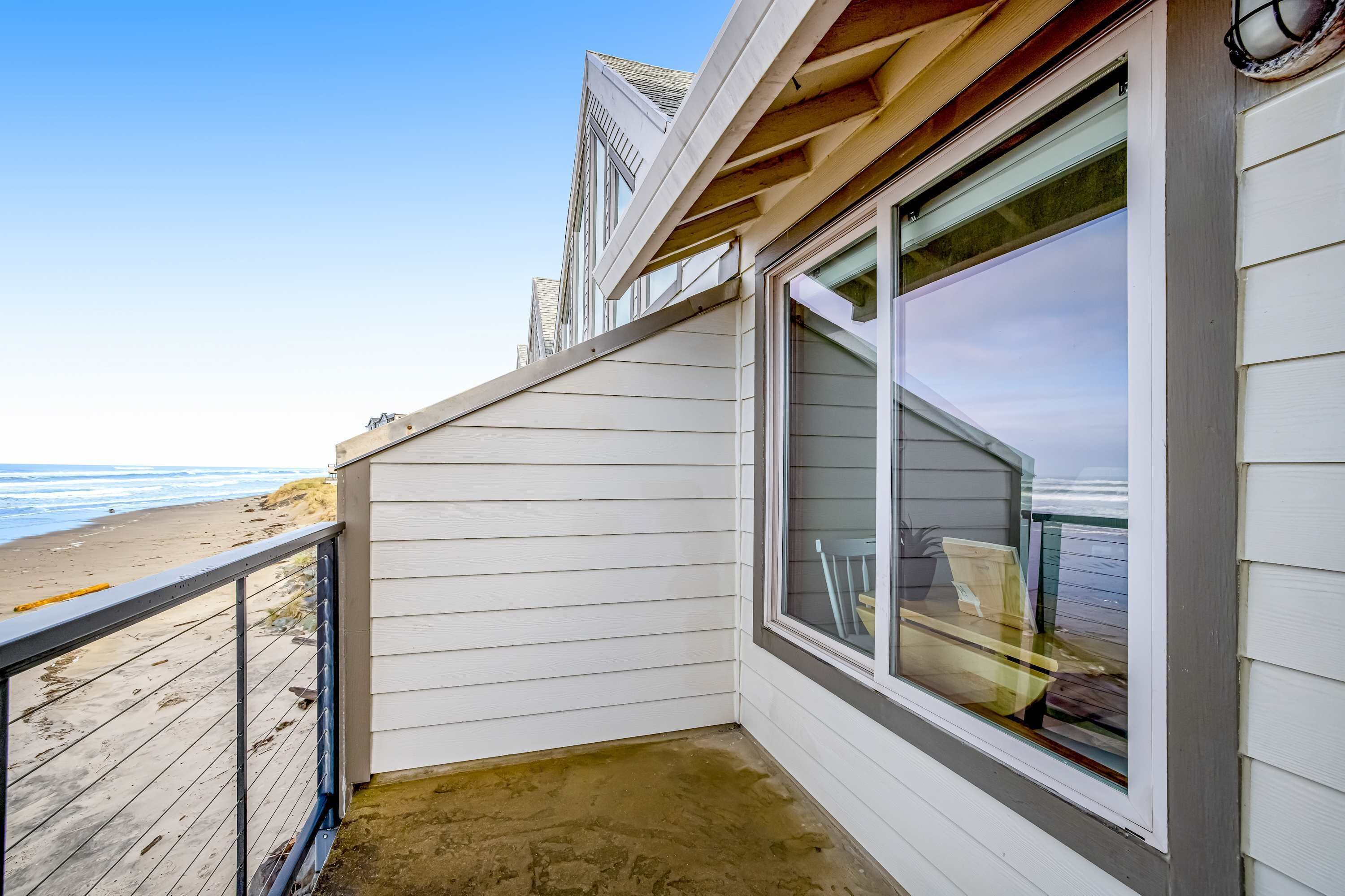 A beach house with a balcony overlooking the ocean, featuring a large window and a wooden deck.