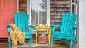 Eine Veranda mit zwei türkisfarbenen Adirondack-Stühlen, einem kleinen gelben Tisch mit Tassen und einem gewebten Korb.