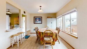 A dining area with a table set for four, wooden chairs, a bar area with stools, a kitchen visible through an open doorway, and a large window providing a view of the outside. - Ocean view home with prime walkable location near beach and town 2 dogs OK (Rockaway Beach)