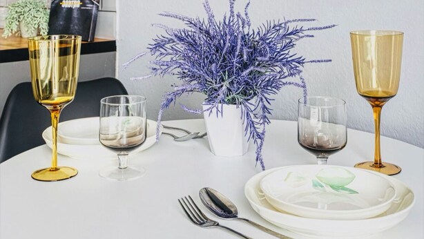 Una mesa de comedor dispuesta con copas de vino, platos y cubiertos, una planta en maceta y un televisor de pantalla plana montado en la pared.