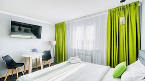 Habitación de hotel con cama, escritorio y sillas, una mesita con planta, y ventana con cortinas translúcidas.