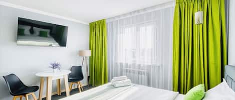 A hotel room with a bed, a desk with chairs, a small table with a plant, and a window with sheer curtains.