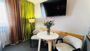 Una habitación de hotel con una mesa redonda, dos sillas, un sofá, un televisor en la pared, una ventana con cortinas y un jarrón con flores sobre la mesa.