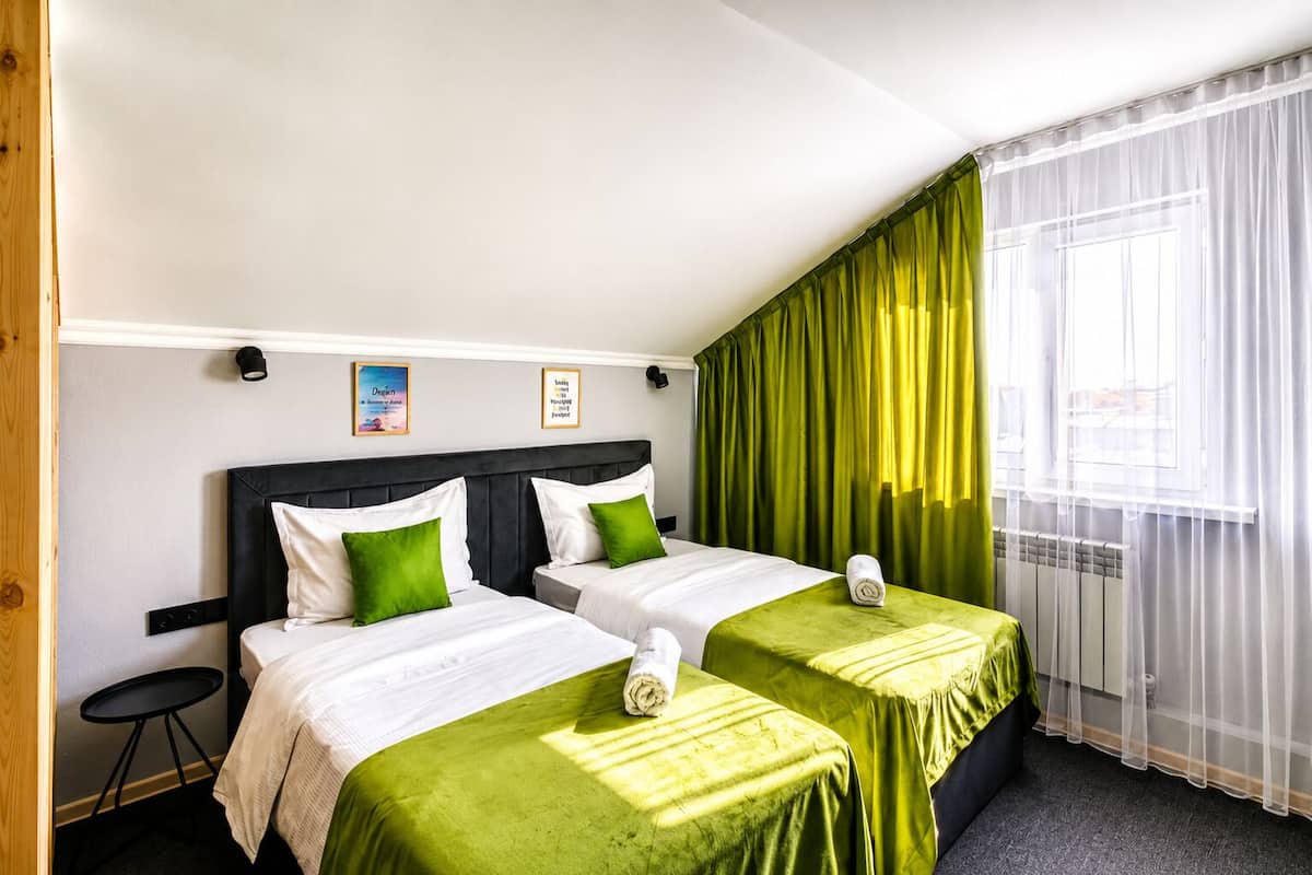 Habitación de hotel con dos camas, cubrecamas verdes, una mesita redonda y una ventana con cortinas translúcidas.