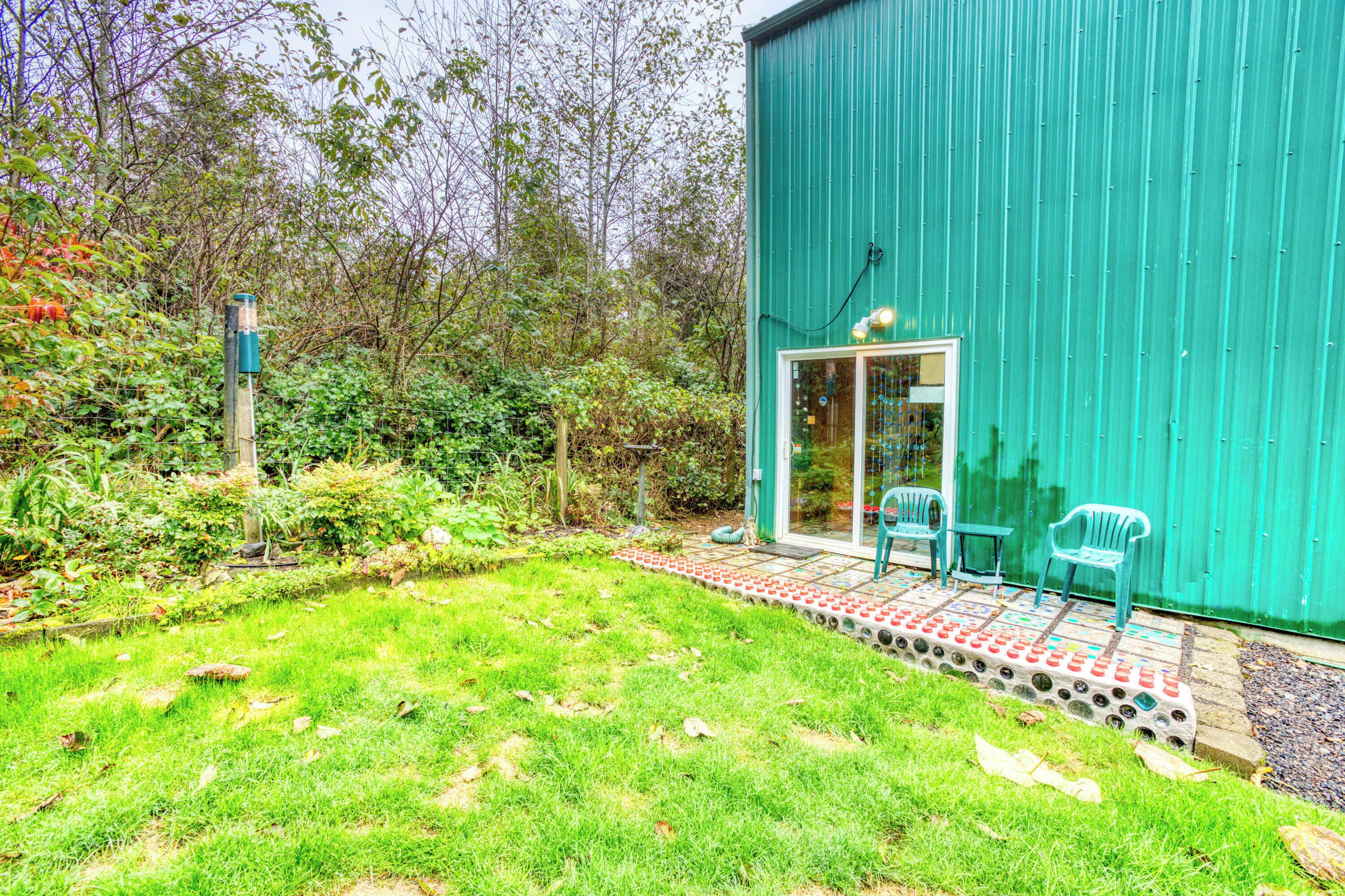 Un bâtiment écologique doté d’une porte et de fenêtres en verre, d’une terrasse pavée avec des chaises, et d’une cour verdoyante avec des arbres et des arbustes.