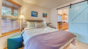 Quarto com cama, mesa de cabeceira, luminária, janela com persianas e porta deslizante para uma cozinha.