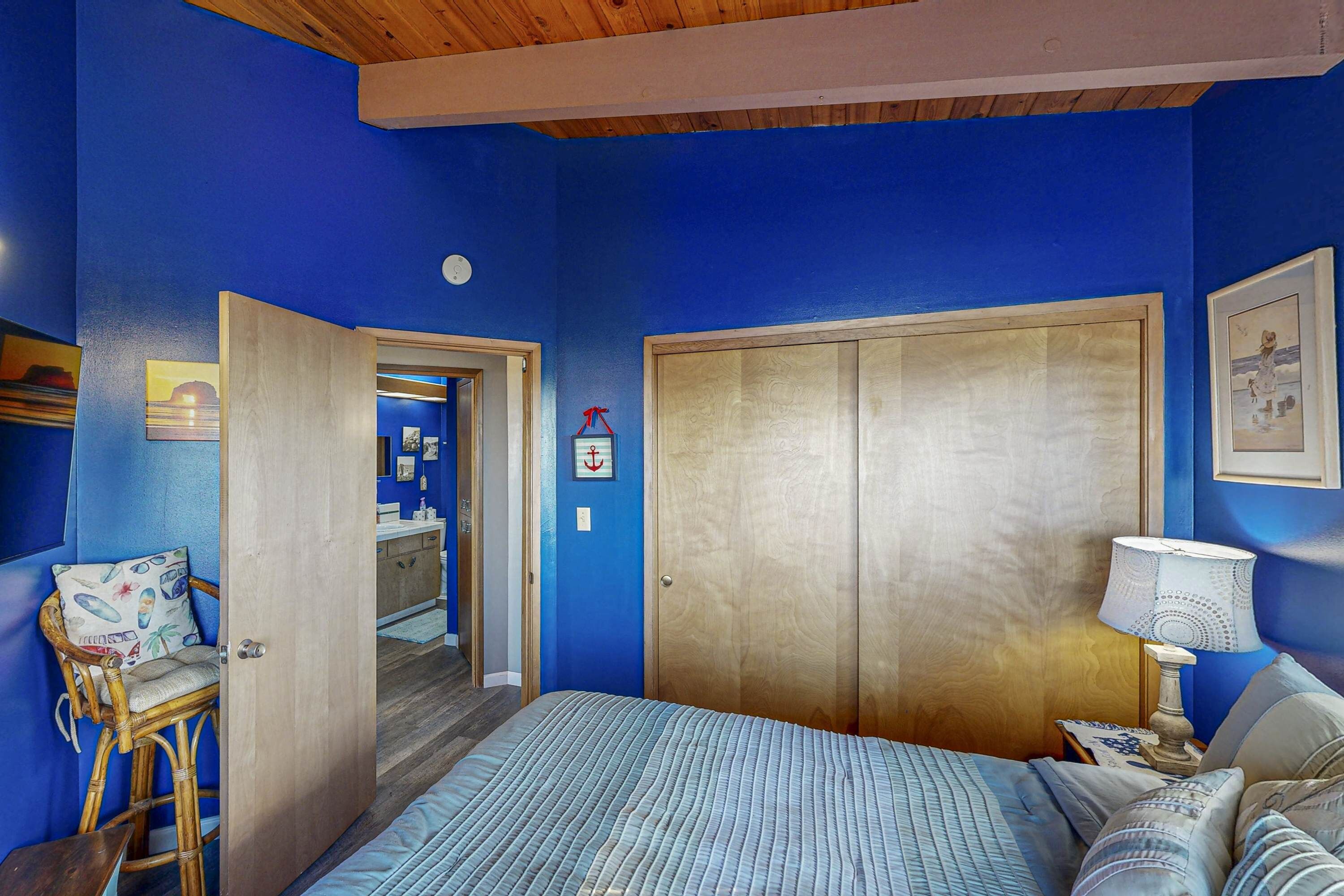 A bedroom with a large bed, a wooden wardrobe, a chair with a cushion, a bedside table with a lamp, and a framed picture on the wall.