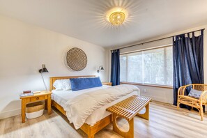Ein Schlafzimmer mit einem hölzernen Bett, einem Nachttisch, einer Bank, einem Korbstuhl und einem großen Fenster mit Vorhängen.