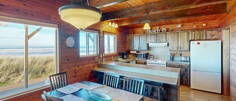 A wooden cabin with a dining area, kitchen, and large windows overlooking the beach.