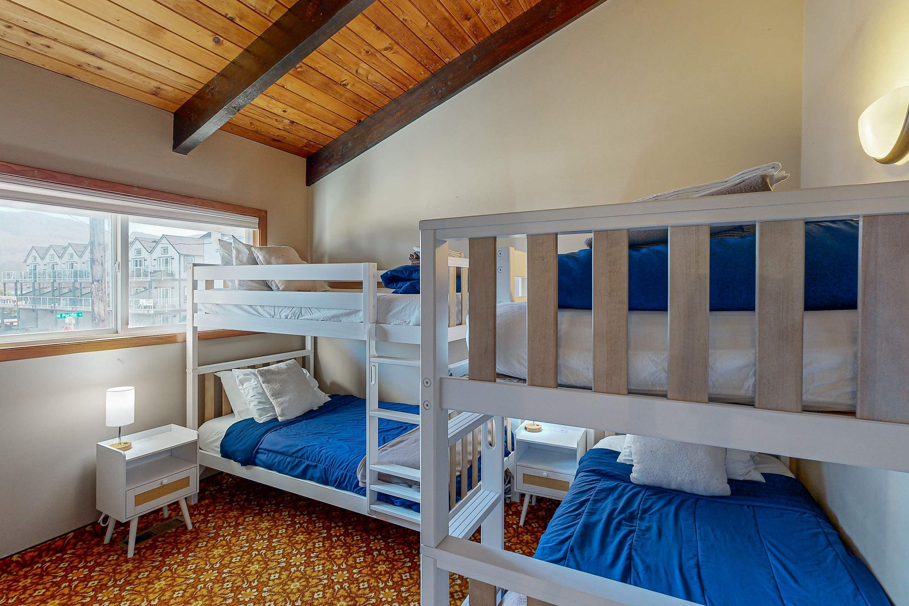 Une chambre avec des lits superposés, une petite table de chevet avec une lampe et une fenêtre donnant sur des immeubles.