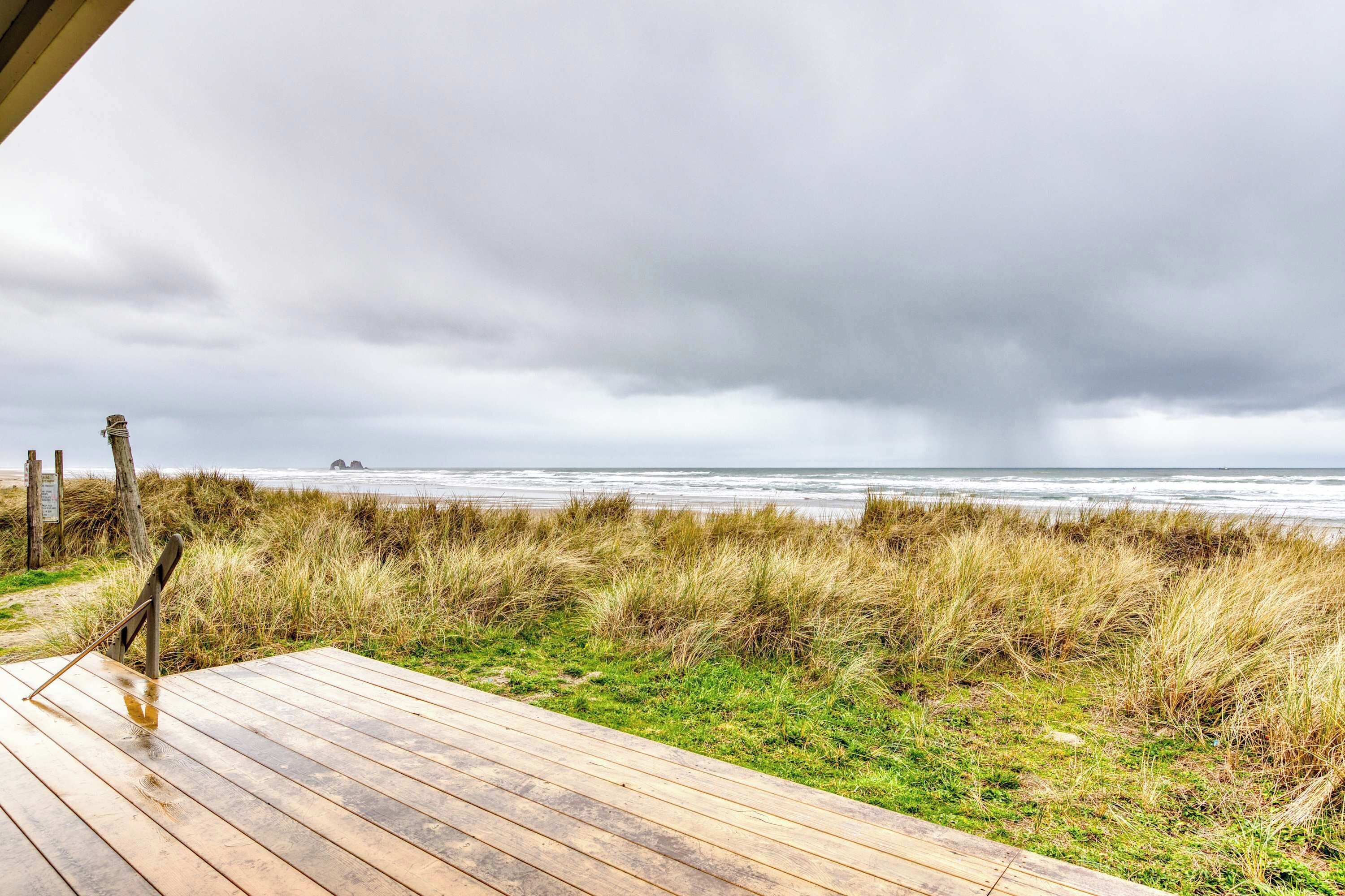 Une terrasse en bois donnant sur une plage, avec des dunes herbeuses et un ciel nuageux.