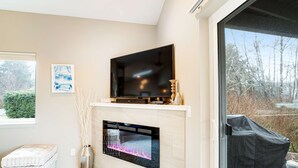 A modern living room with a flat-screen TV mounted above a fireplace, a sofa, and a window with a view of trees. - New Family Friendly Home with a Furnished Patio and Tranquil Yard (Rockaway Beach)