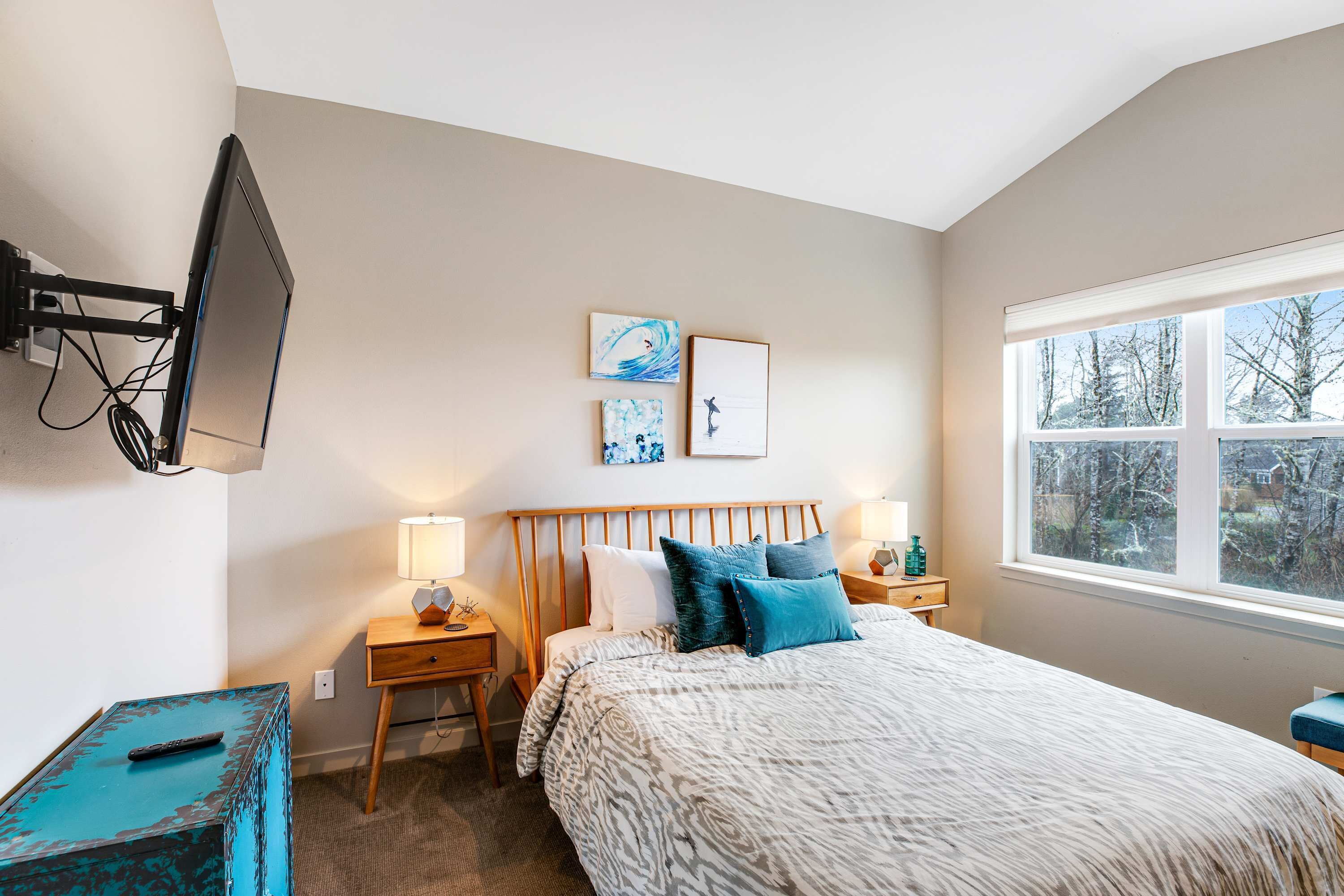 A bedroom with a bed, bedside tables, a TV mounted on the wall, and a window with a view of trees.
