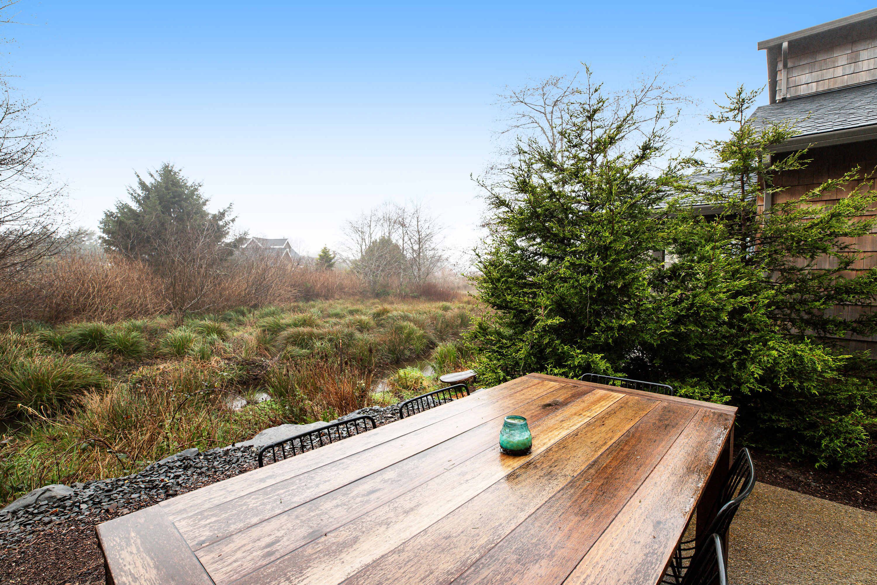 An outdoor wooden table with a green vase, surrounded by greenery and a stone pathway.