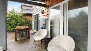 A patio with a wooden table and chairs, a wicker chair, and a sliding glass door leading to a covered area. - New Family Friendly Home with a Furnished Patio and Tranquil Yard (Rockaway Beach)