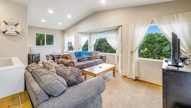 Ein geräumiges Wohnzimmer mit grauem Sofa, einem Couchtisch und einem Fernseher auf einem Schrank. Große Fenster mit Vorhängen, eine Wanduhr und ein Kamin sind ebenfalls vorhanden.