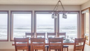 Un comedor con mesa dispuesta para seis personas, ventanas amplias con vistas al océano y una lámpara colgante.