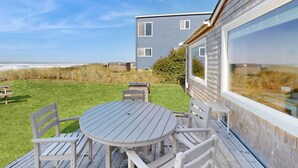 A deck with a table and chairs, overlooking a beach and a building with large windows. - Quaint Beach Getaway with Beach Access Ocean View and Perfect Location Near Town (Rockaway Beach)