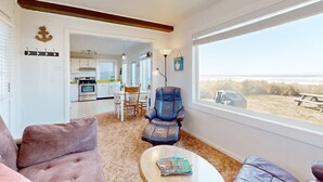 A living room with a sofa, a coffee table with magazines, a blue chair, and a view of the beach through a large window. - Quaint Beach Getaway with Beach Access Ocean View and Perfect Location Near Town (Rockaway Beach)