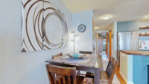 Een eetgedeelte met een tafel voor vier personen, een groot abstract schilderij aan de muur en een keuken met een koelkast en kasten.