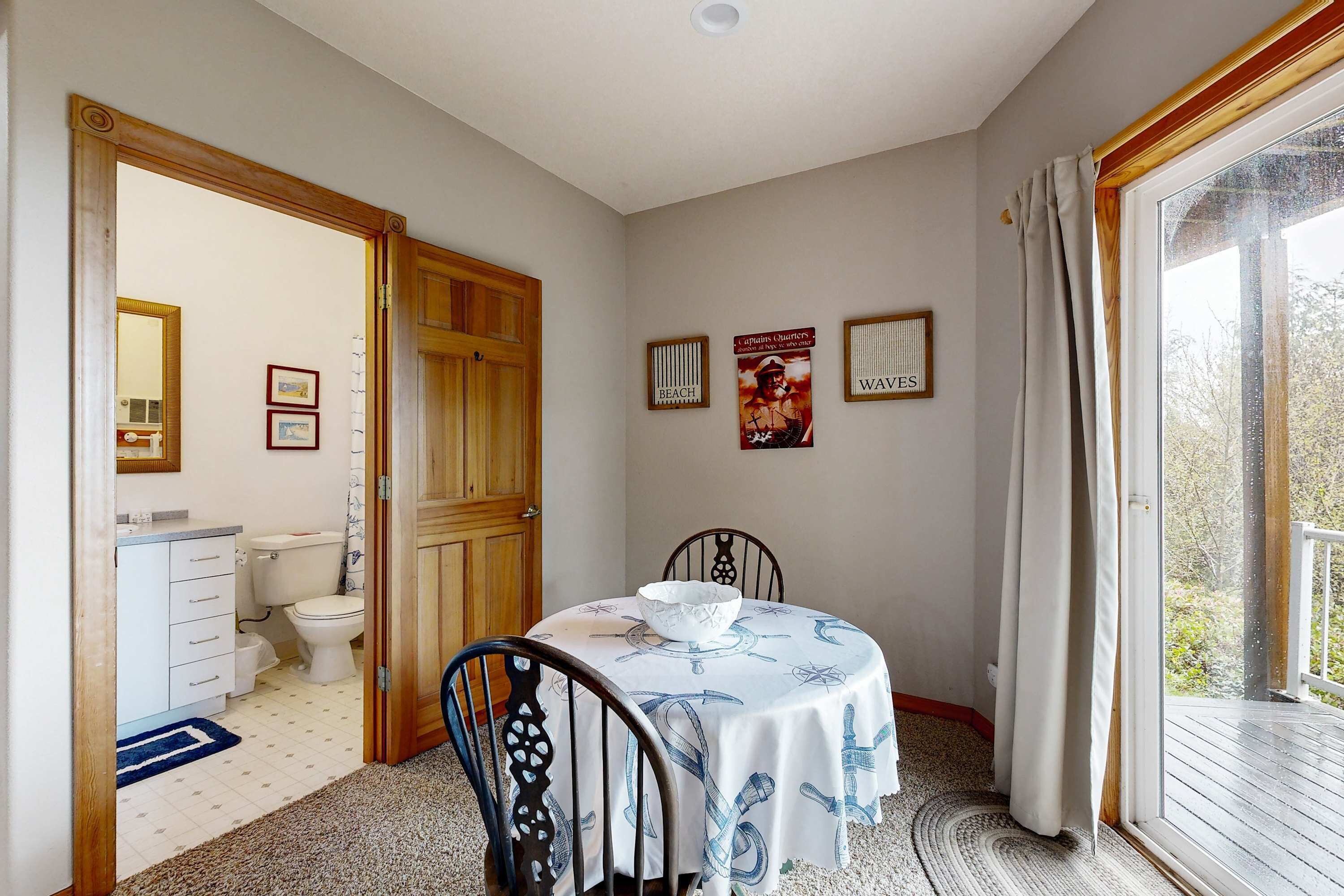 A room with a dining table, chairs, a window with curtains, a wooden door, and a bathroom visible through an open door.