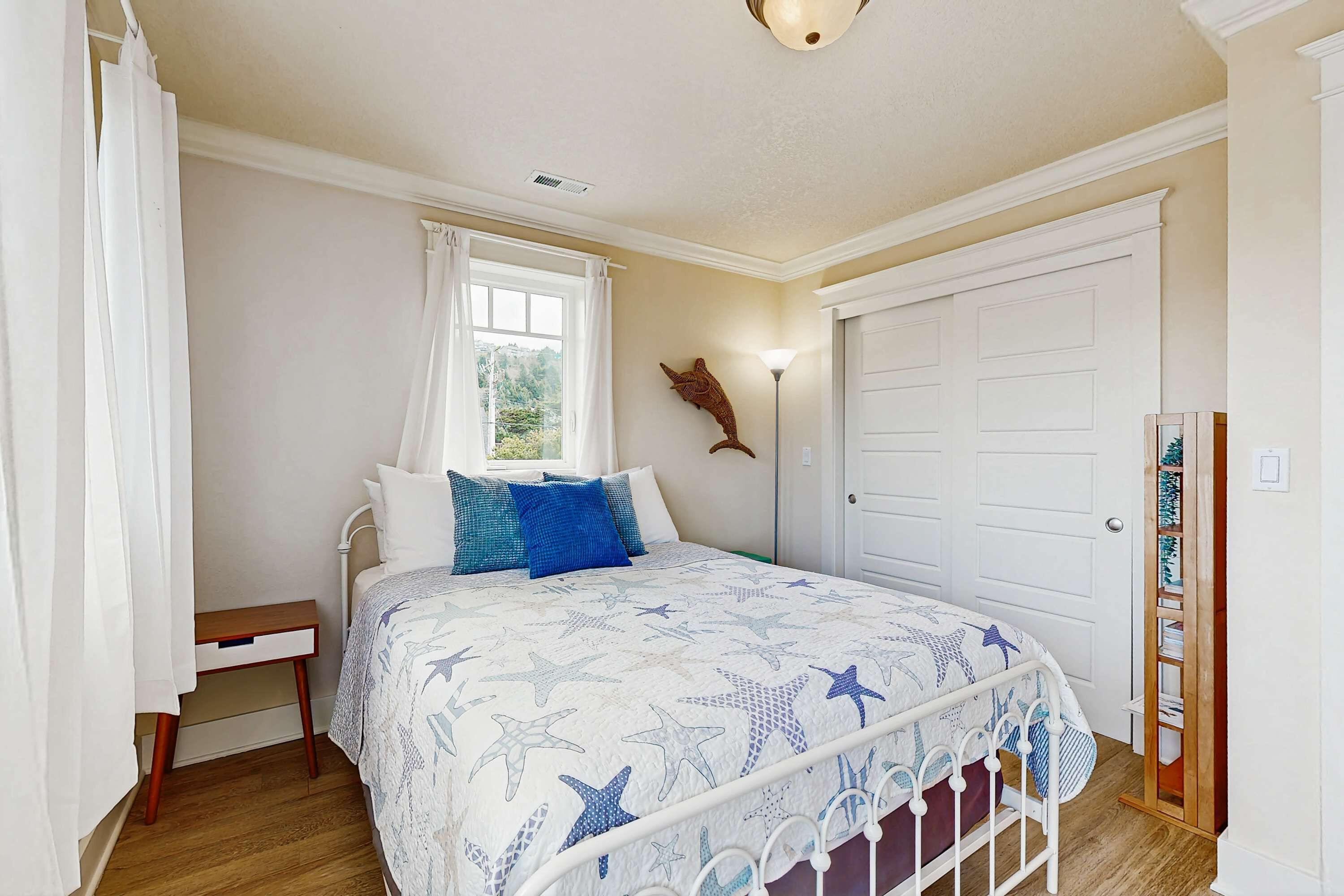 Un dormitorio con cama, mesita de noche, ventana con cortinas, lámpara de pared y puerta.