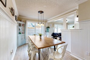 A dining area with a wooden table and white chairs, a light blue cabinet, and a ceiling light fixture. - Dog friendly beach home with firepit private hot tub beach access and arcade (Rockaway Beach)