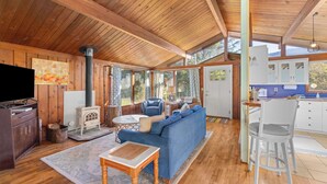 A cozy cabin interior with a wooden ceiling, a blue sofa, a dining area, and a kitchen with white cabinets.