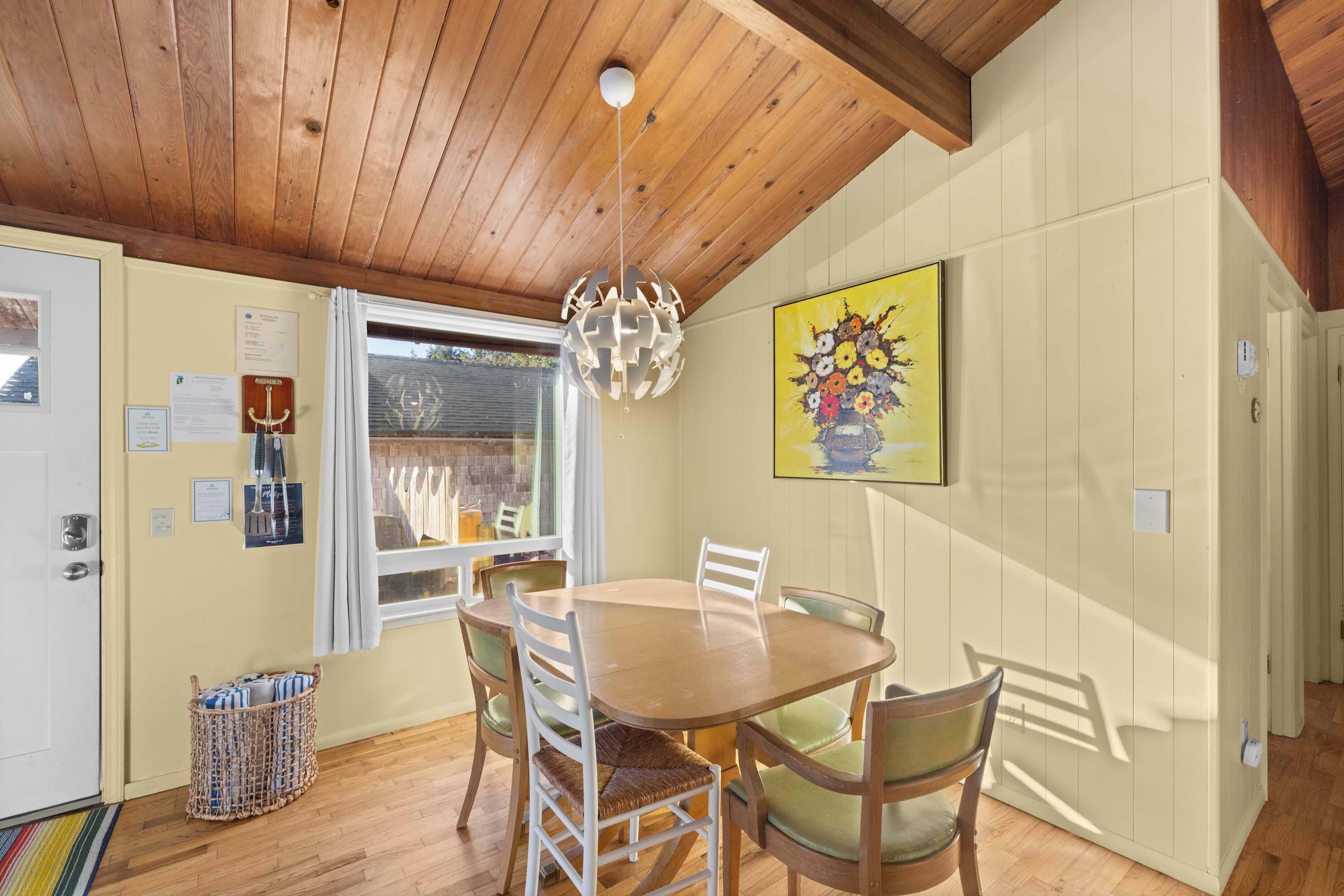 A dining area with a wooden table and chairs, a hanging light fixture, a window with curtains, and a painting on the wall.