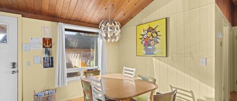 A dining area with a wooden table and chairs, a hanging light fixture, a window with curtains, and a painting on the wall.