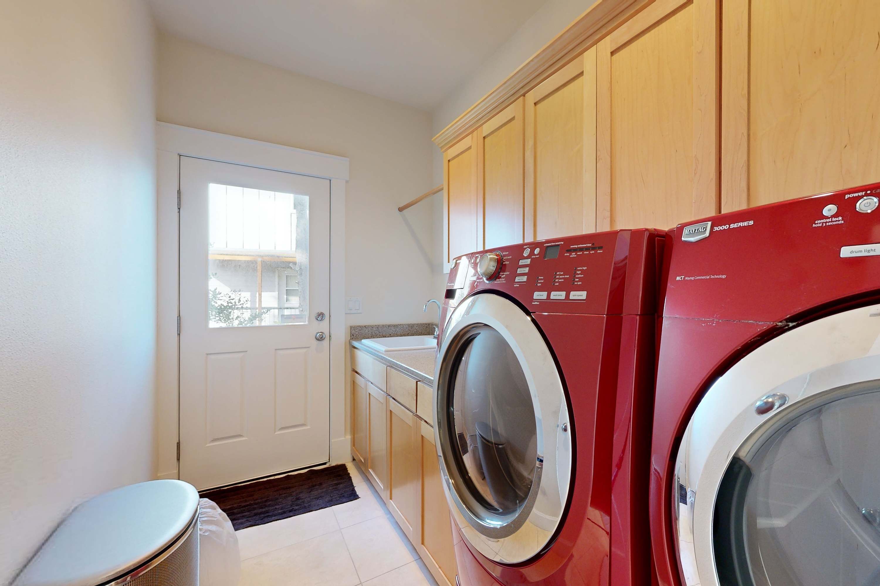 A laundry room with a red washing machine, a white dryer, wooden cabinets, and a white door with a window.