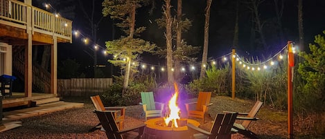 An outdoor seating area with a fire pit surrounded by chairs, string lights, and a deck.