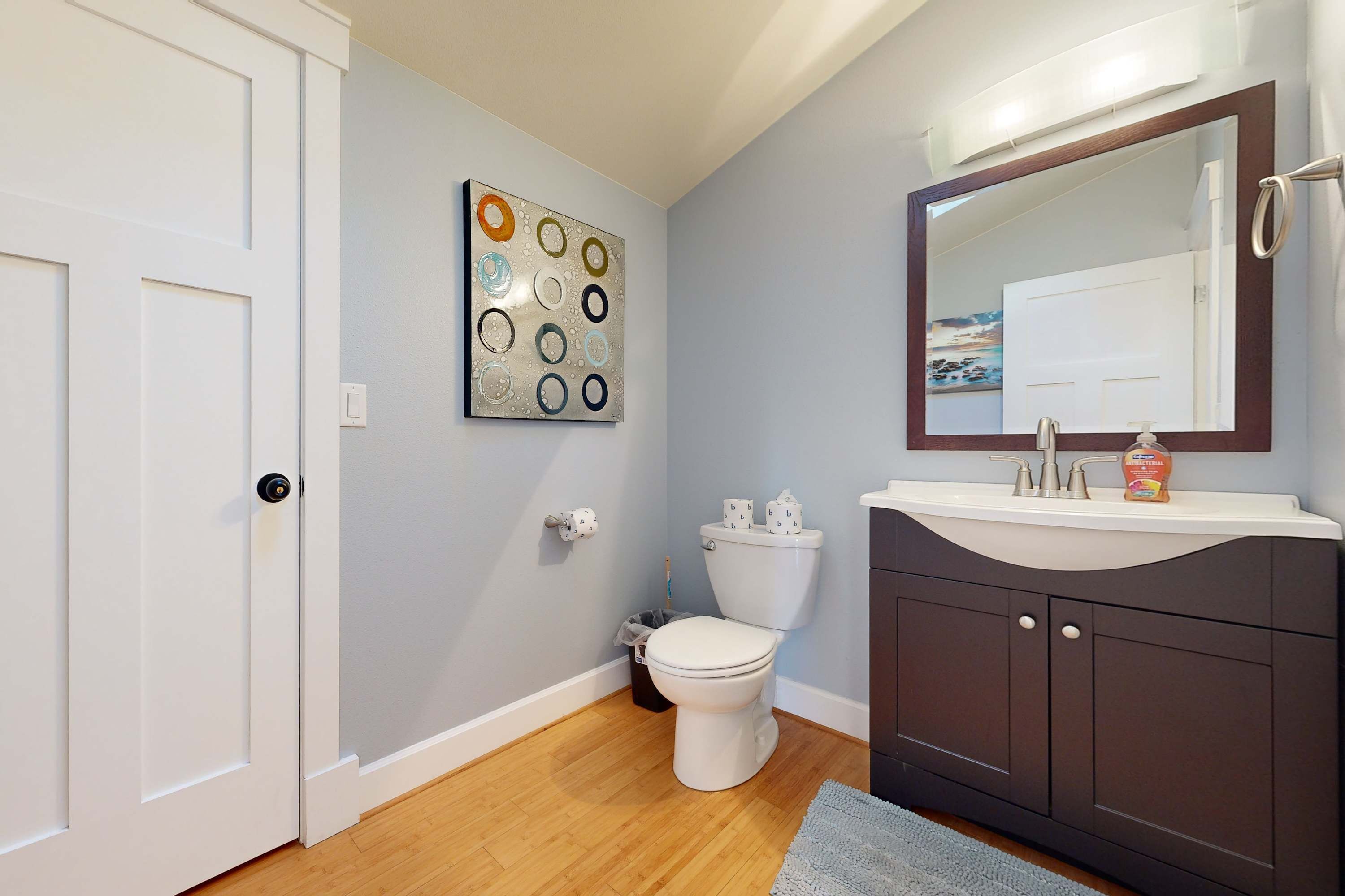 A bathroom with a white toilet, a dark vanity, a mirror, and a wall art piece.