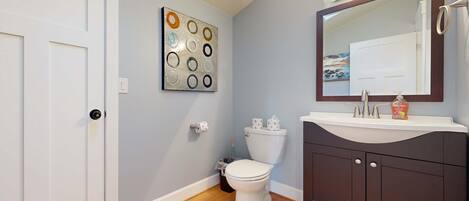 A bathroom with a white toilet, a dark vanity, a mirror, and a wall art piece.