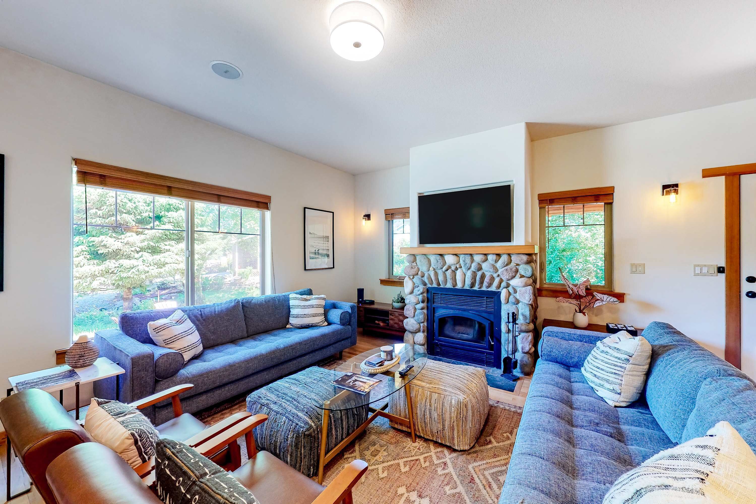 A cozy living room with a stone fireplace, blue sofas, and a large window offering a view of greenery.