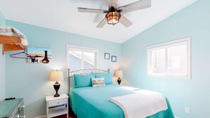 Una camera da letto con una coperta color acquamarina, un ventilatore a soffitto, un comodino con lampade, un comò e una finestra con persiane.