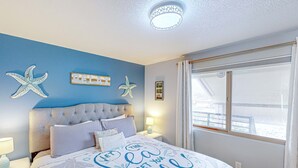 Une chambre avec un mur bleu, des décorations en forme d’étoiles de mer, un lit recouvert d’une courtepointe sur laquelle est inscrit « Laissez la mer vous libérer », une table de chevet avec une lampe et une fenêtre avec des rideaux.