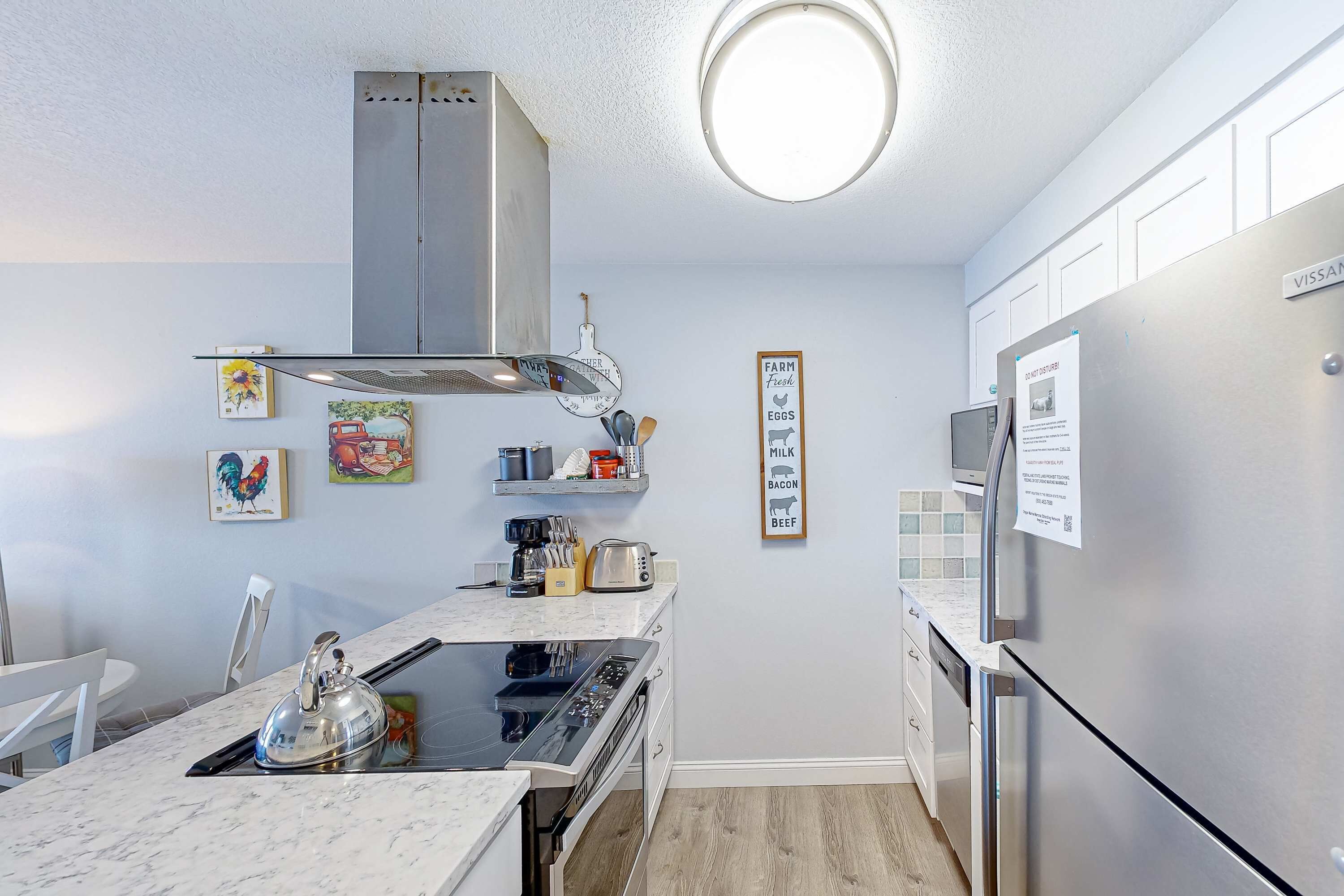 A compact kitchen with a stainless steel range hood, a built-in oven, a microwave, and a refrigerator. There are wall-mounted shelves with kitchen utensils and a framed sign listing farm-fresh eggs, milk, bacon, and beef.