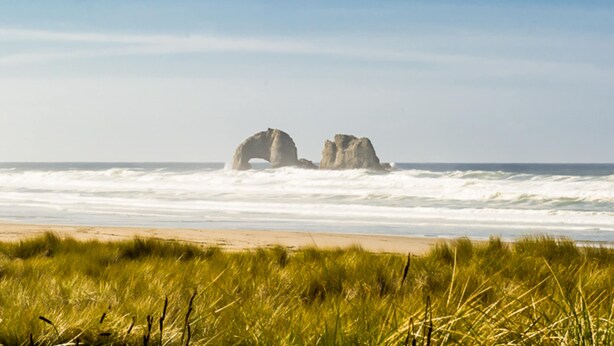 Ein Strand mit zwei großen Felsen im Hintergrund, einem sandigen Ufer und Dünen im Vordergrund.