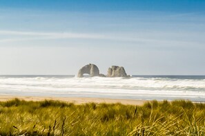 A beach with two large rock formations in the distance, a sandy shore, and grassy dunes in the foreground. - Ocean view townhouse 100 ft to beach with fireplace Ping Pong and private garage (Rockaway Beach)