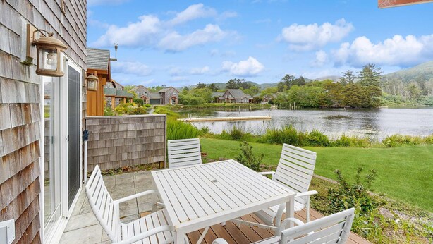 Una terraza de madera con mesa y sillas da vista a un lago, rodeada de follaje y otros edificios.
