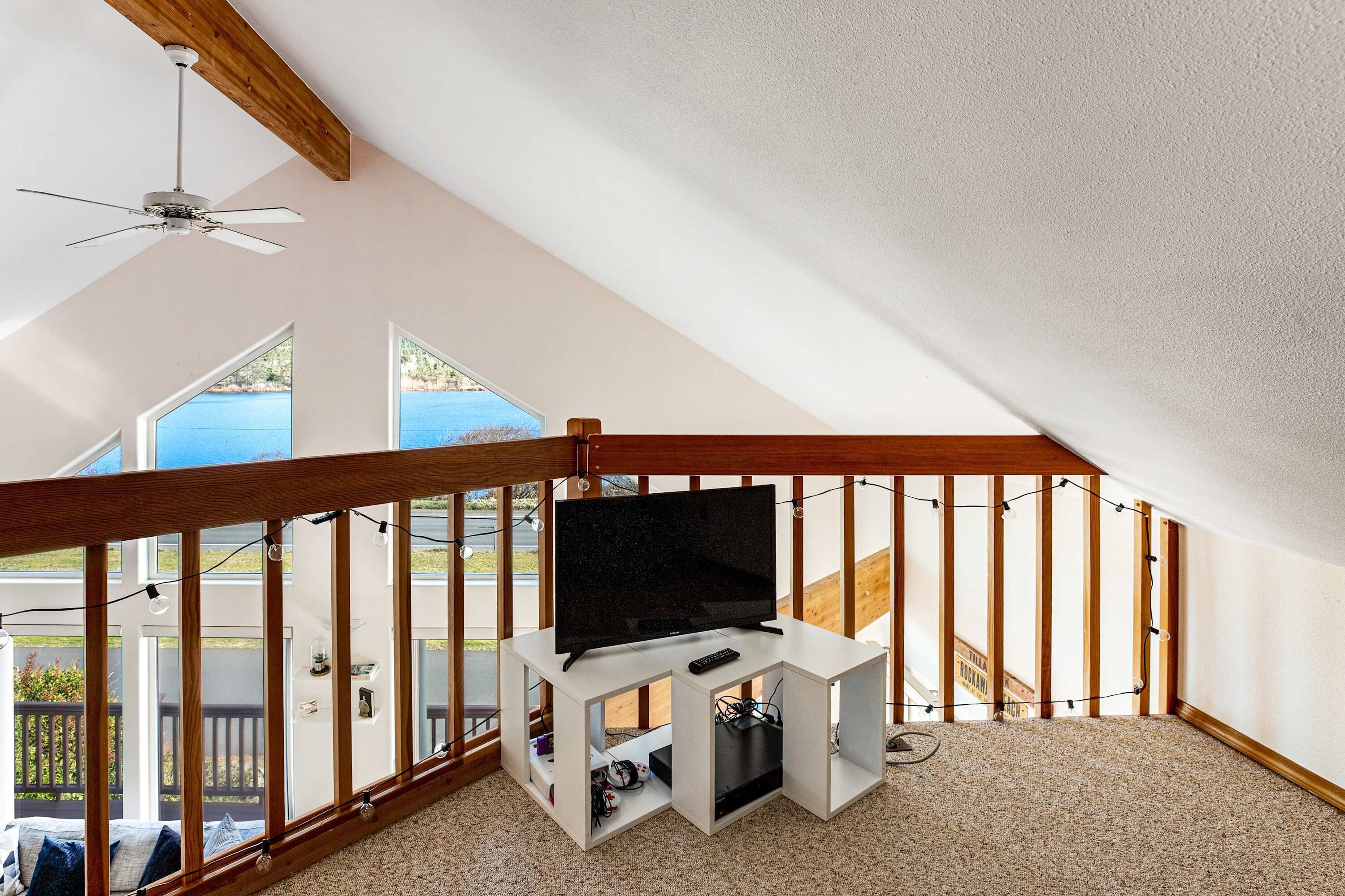 Un salon moderne avec un téléviseur à écran plat posé sur un meuble de télévision blanc, un ventilateur de plafond et de grandes fenêtres donnant sur l’extérieur.