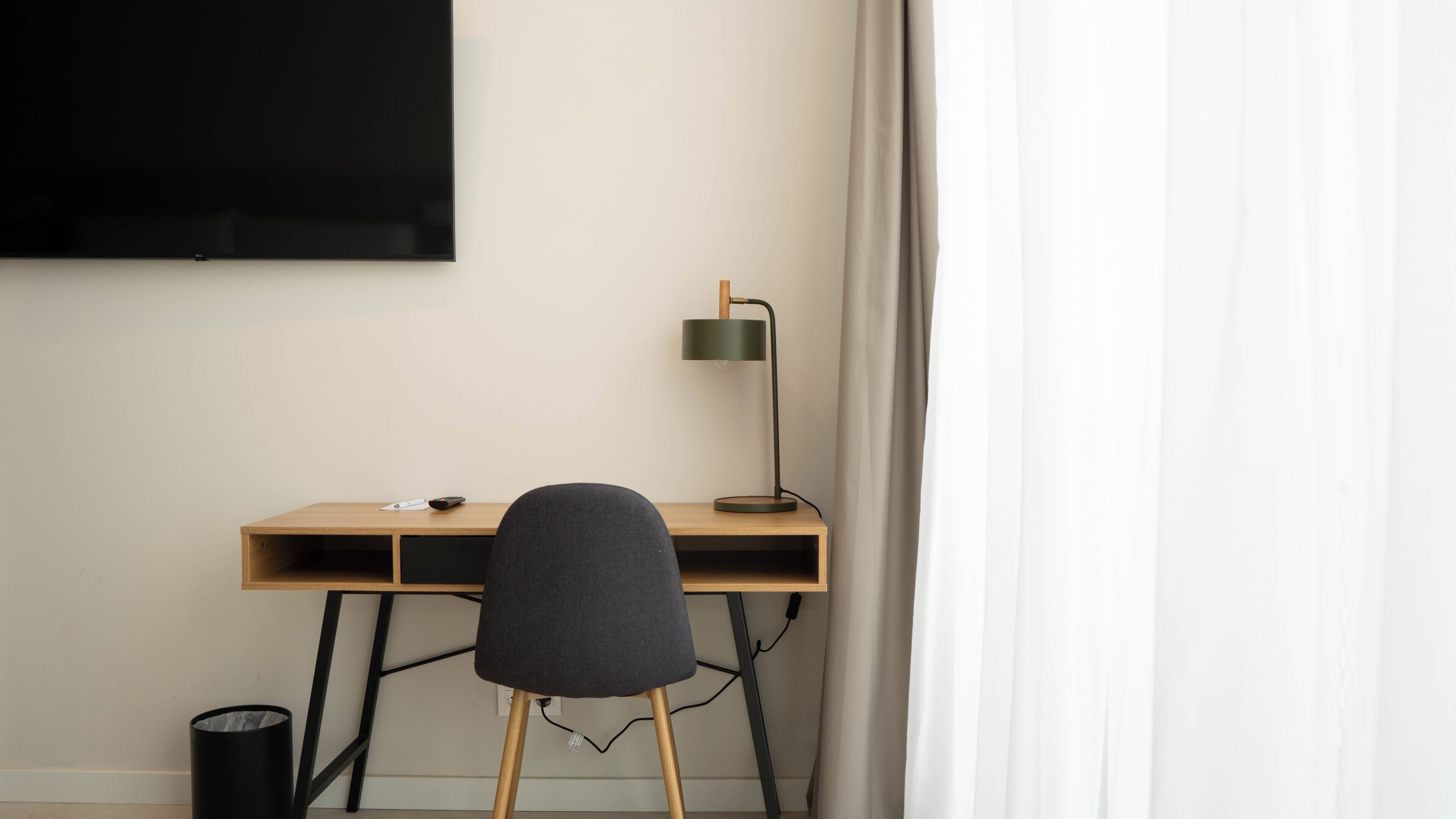 A minimalist workspace with a wooden desk, a black chair, a floor lamp, and a black trash can.
